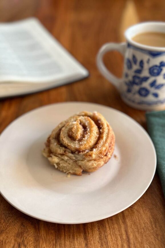 single plated cinnamon roll on white plate with a coffee mug napkin and open bible