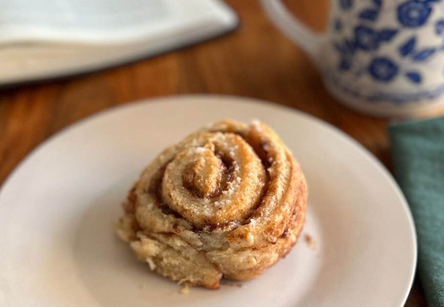 single plated cinnamon roll on white plate with a coffee mug napkin and open bible