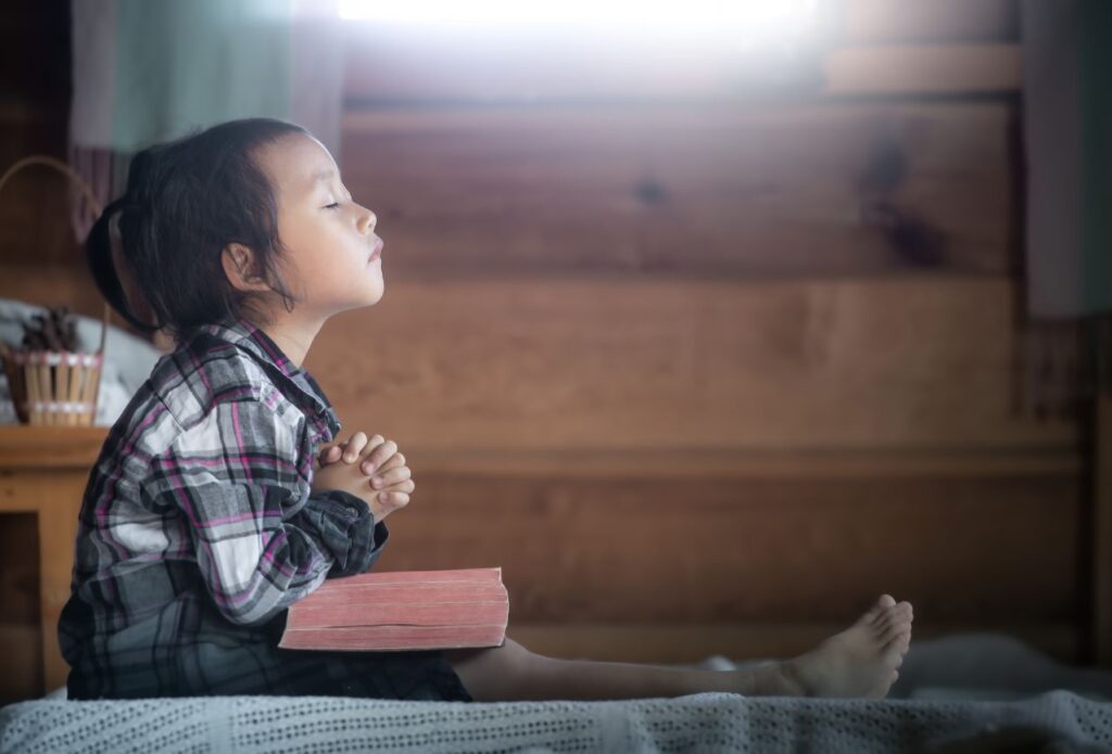 small child seated in room with hands folded in prayer and a bible in her lap