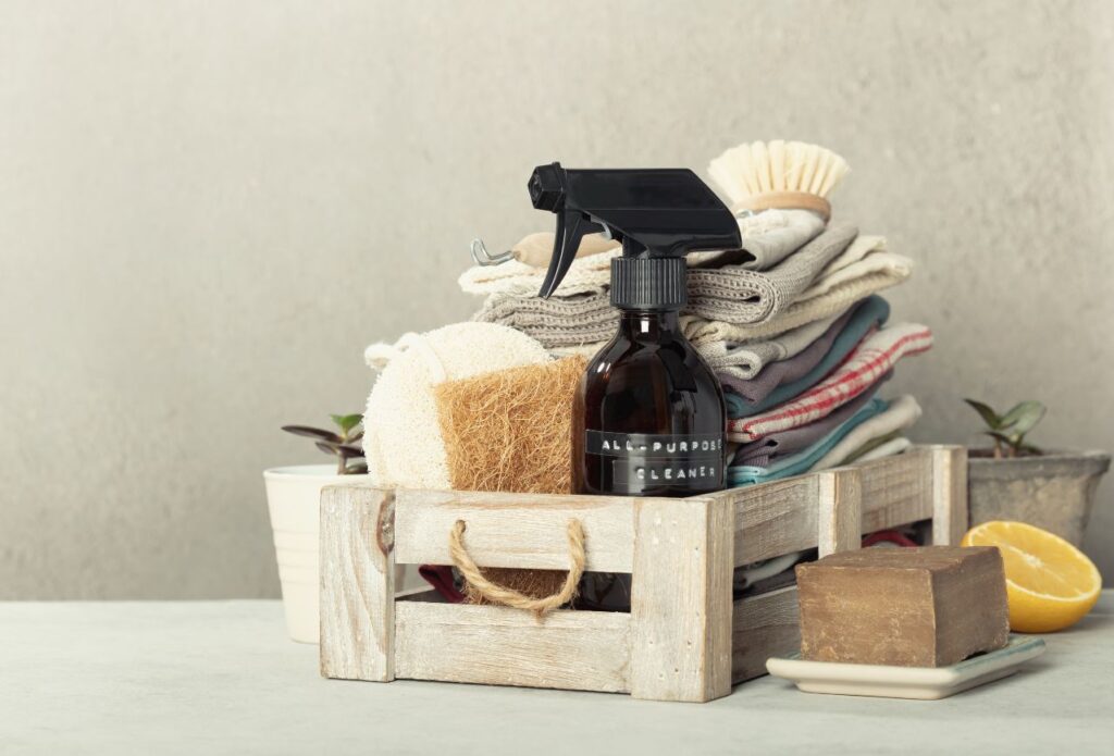 dark spray bottle of diy cleaner in a basket with natural cloths, loofa, bar of soap and scrub brush