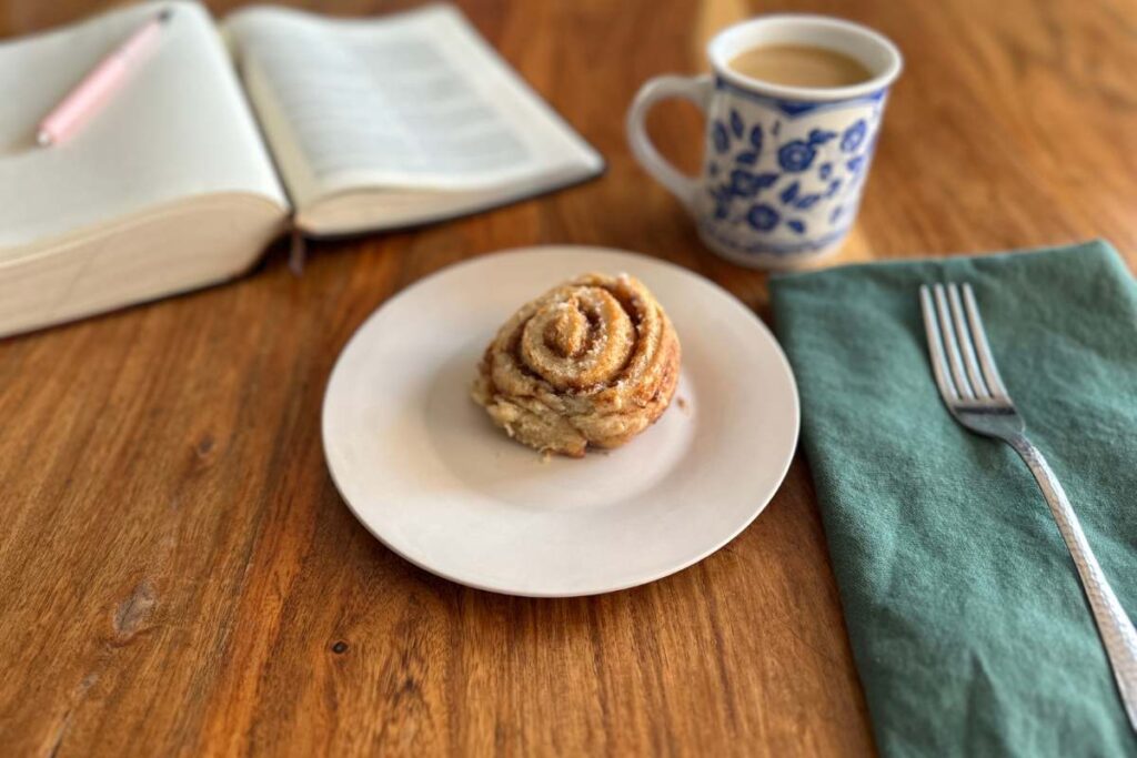 plated cinnamon roll with fork and napkin, coffee cup, and open bible