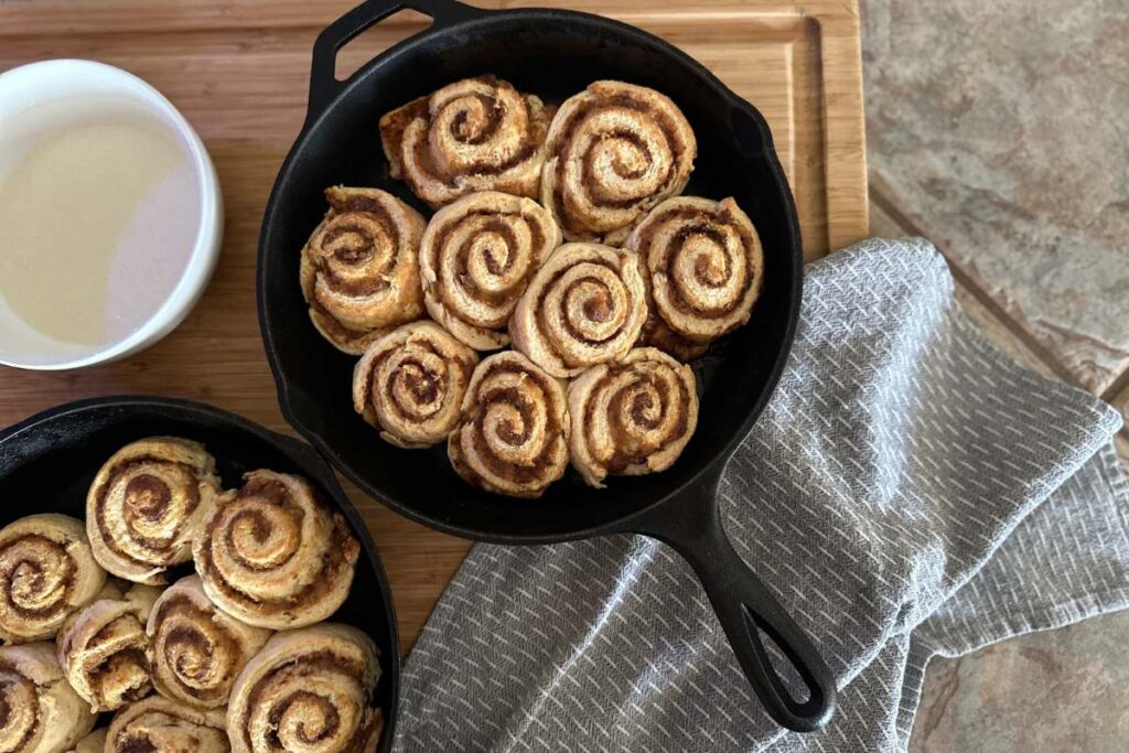 two skillets of baked cinnamon rolls with a small bowl of mixed glaze and a blue towel